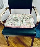 Wooden chair with black faux leather seat and back, floral cushioned chair pad on seat, wooden arm rests, placed on hardwood floor.