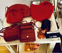 Photo showing a variety of red handbags and purses on a small white table, including quilted and fur-style bags, with a small black wallet labeled 'Las Vegas'.