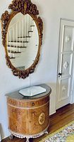 Ornate gilt mirror hanging above a half-moon inlaid wood credenza with marble top