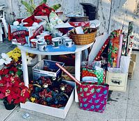 White round table with various Christmas decorations including mugs, bowls, poinsettias, bows, ornaments, and ribbons. Box under table contains gold accented holiday ornaments. Behind table are multiple rolls of Christmas wrapping paper and gift bags.