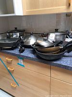 Photo showing various cookware items including slow cooker, electric skillet, frying pans, and wok on a kitchen counter.