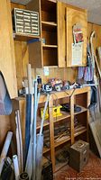 Wide shot of basement with wood shelving, various tools scattered including jigsaw, hand saw, soldering tool, and wood scraps.