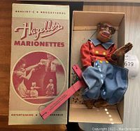 Photo showing the Hazelle's Marionette Jocko the Monkey puppet lying in original box with the box lid propped beside it. Puppet wears red jacket and blue pants, strings and control bar attached. Shows flaking and some damage.