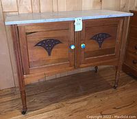 Front view of wooden cabinet showing two panelled doors with carved decorations and colorful knobs, with marble top visible, mounted on caster wheels.