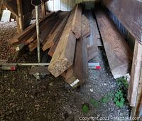Various sizes and types of lumber stacked on a gravel ground beneath a shelter, includes wide and narrow pieces.