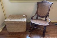 Wider view showing carved wooden rocking chair with woven wicker seat and square woven wicker basket beside it on wooden floor.