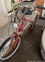 Front left angle view showing orange front fork, black handlebar grips, black padded seat and metal wire basket over rear wheels.