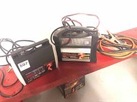 Photo showing two battery chargers and jumper cables on a red surface.