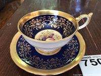 Photo showing side view of teacup and saucer with gold rim, blue background, gold floral detailing, and painted fruit center.