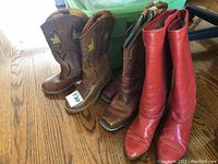 View of three pairs of boots lined up on a hardwood floor including red leather knee-high boots and two pairs of cowboy boots with decorative stitching.