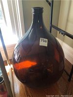 Photo of a large brown glass carboy bottle on a wooden surface beside a window with natural light showing its amber translucent color and narrow neck with rounded body.