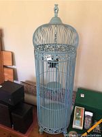 Full view of the tall decorative light blue iron bird cage with domed top and finial, placed on floor with other items nearby.