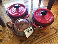 Photo showing two red Le Creuset covered casseroles and one brown ceramic teapot on a wooden surface.