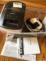 Overall view of the Brother QL-700 label printer sitting in a clear plastic bin with the roll of label tape and manuals.