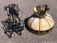 Iron chandelier and slag glass pendant lamp placed side by side outdoors on asphalt surface. Chandelier is black wrought iron with leaf and teardrop design; pendant lamp has segmented cream and amber slag glass with bronze decorative trim.