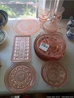 Photo of various pink depression glassware items including plates, serving tray, goblet, and small bowls showing intricate starburst and geometric patterns.