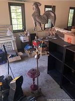 Full view of the rooster weathervane mounted on a tall stand in a garage setting surrounded by storage items and shelving units.