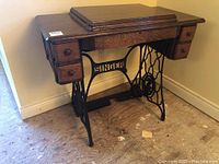 Full view of antique Singer sewing machine cabinet with iron base and multiple small drawers, closed tabletop and visible signs of wear on wooden surface.