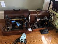 Two vintage Singer sewing machines placed side by side on wooden floor, both with wooden carrying cases and foot pedals.