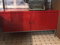 Red steel two door cabinet angled view showing key lock handles and ventilation slots on doors, sitting on metal legs on tiled floor.