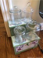 Photo showing two-step decorative display with floral painted top and front with four clear glass apothecary jars with lids, one jar includes a scoop.