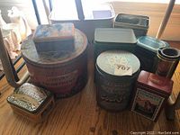 Overview of vintage storage tins showing various branded and decorated metal tins of different shapes and sizes arranged on wooden floor.
