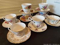 Overview of all 8 bone china teacups and saucers arranged on a black surface showing the variety of floral patterns and gold trim