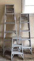 Three aluminum step ladders positioned side by side against a wall with a window. The tallest is a 5-step ladder, the middle a 4-step, and the shortest a 2-step ladder.