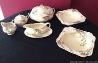 Full set of antique Crown Duval Horentine Rosalie porcelain serving pieces arranged on black cloth, showing all items in the lot including covered serving dish, two squared bowls, gravy boat, cream and sugar set, and platter.