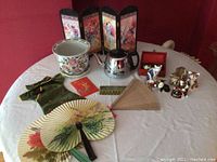 Wide view of the lot on a white table cloth showing plant pot, metal tea pot, folding fans, table divider, wine bottle cover, and cat figurines