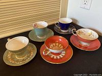 Five Aynsley bone china teacups and saucers in various pastel and deep colors with gold embellishments arranged on a black surface.