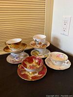 Six bone china teacups and matching saucers displayed together on a black surface, showing a variety of floral patterns and gold trim detailing.