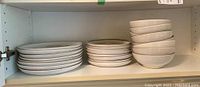Three stacks of off-white Denby stoneware on shelf: dinner plates on left, salad plates in middle, soup bowls on right.