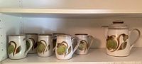 Six Denby Troubadour mugs and one coffee pot on a white shelf. Items show the green bell-shaped floral design and brown accents on white stoneware.
