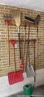 Photo of gardening and outdoor tools hung on a brick wall, showing two red snow shovels, various garden hand tools including spade, fork, cultivator, a corn broom, push broom and green watering can on the floor.