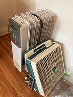Radiator heater standing upright showing metal fins and control panel. Beige and teal Super brand floor fan leaning against heater.