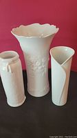 Three white vases against a red background: large scalloped rim porcelain vase with floral embossing in center, plain rounded vase with imprinted basket detail, narrow modern folded white vase