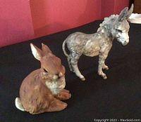Photo showing both the porcelain donkey and rabbit figurines together on a black surface against a red wall background for overall view.