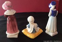 Back view of three Royal Doulton Kate Greenway Collection porcelain figurines against red background showing Sophie, Ellen, and Lucy.