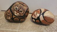 Two large painted rocks showing realistic cats, one with two tabby cats and the other with a calico cat curled up.