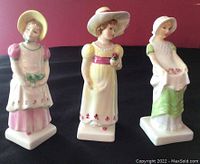 Front view of the three Royal Doulton Kate Greenway figurines positioned side by side on a black surface with red background.