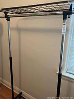 Full view of the collapsible clothing rack showing chrome vertical poles with black plastic connectors and top metal grid shelf.