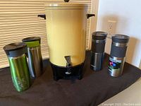 Yellow Westbend percolator with black handles and four assorted stainless steel Contigo travel mugs of different colors and lid designs.