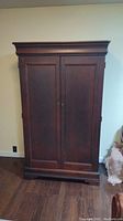 Front view of the solid dark wood armoire with closed paneled doors and brass-colored knobs, showing the entire piece against the wall.