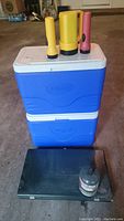 Two stacked blue Coleman coolers with three flashlights placed on top, and Coleman camp stove with propane bottle in front.