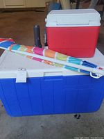 Blue Rubbermaid cooler at the bottom with white lid, red Coleman cooler on top, black flashlight standing on the white lid, and folded multicolored beach umbrella lying across both coolers.