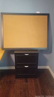 Front view of the framed cork board resting on top of the two drawer file cabinet.