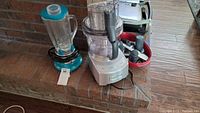 Photo showing Cuisinart blender with turquoise base and glass carafe and the white Cuisinart food processor with clear plastic container and various accessories in a red tray.