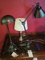 Three desk lamps displayed together showing different designs and power cords, one in black metal with dome shade, one white shade with silver base, one black adjustable arm style.