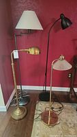 Four floor lamps positioned against a red wall, varying in height from approximately 36 to 60 inches. Shows details of lamp shapes and finishes including brass and black metal.